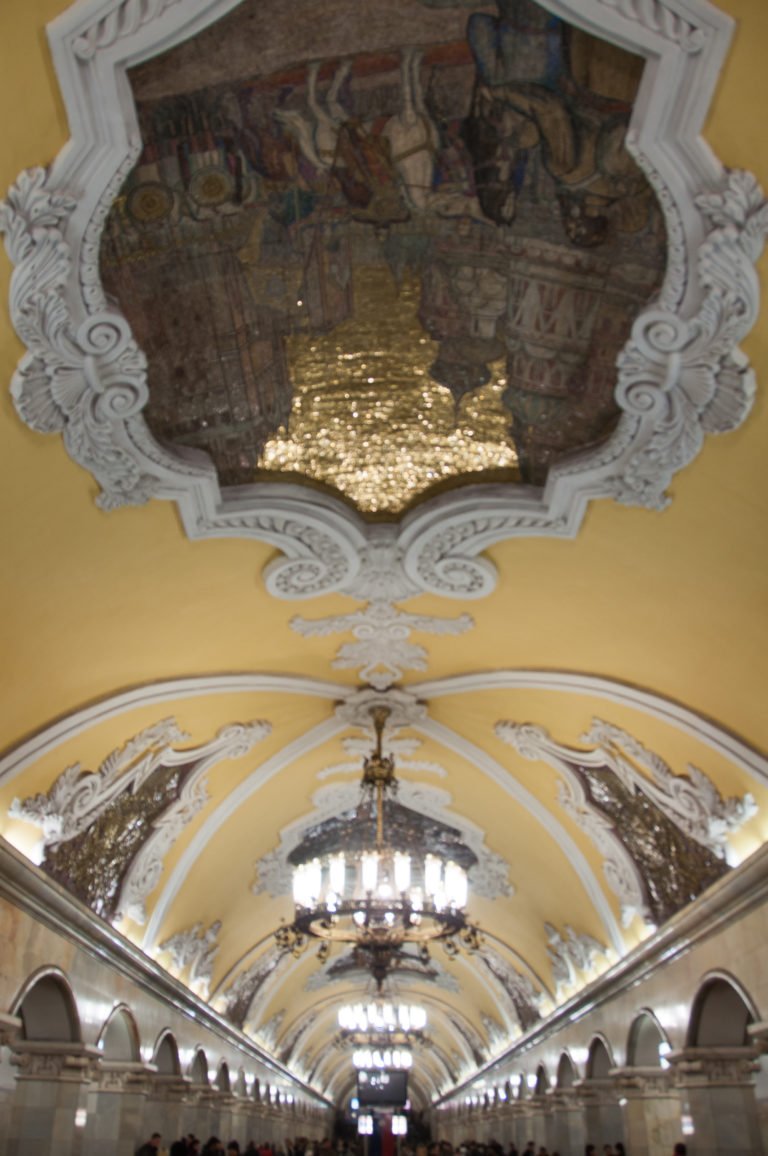 Long yellow vaulted ceiling with repeated chandeliers, white plaster ornament, and framed narrative mosaics receding into the distance.