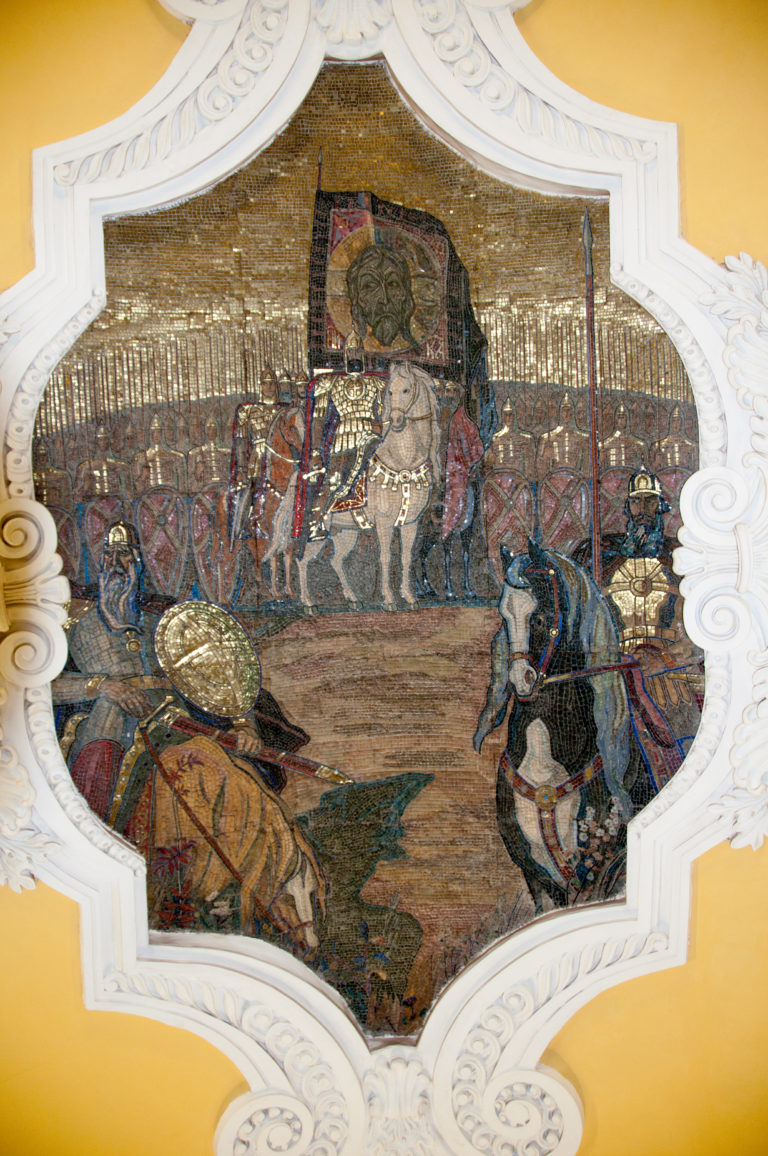 Historical mosaic of mounted figures, banners, and a procession set within an ornate white plaster frame on a yellow ceiling.