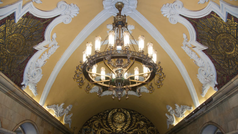 Large chandelier hanging from a yellow vaulted ceiling with white plaster scrolls and dark Soviet-themed mosaic panels.