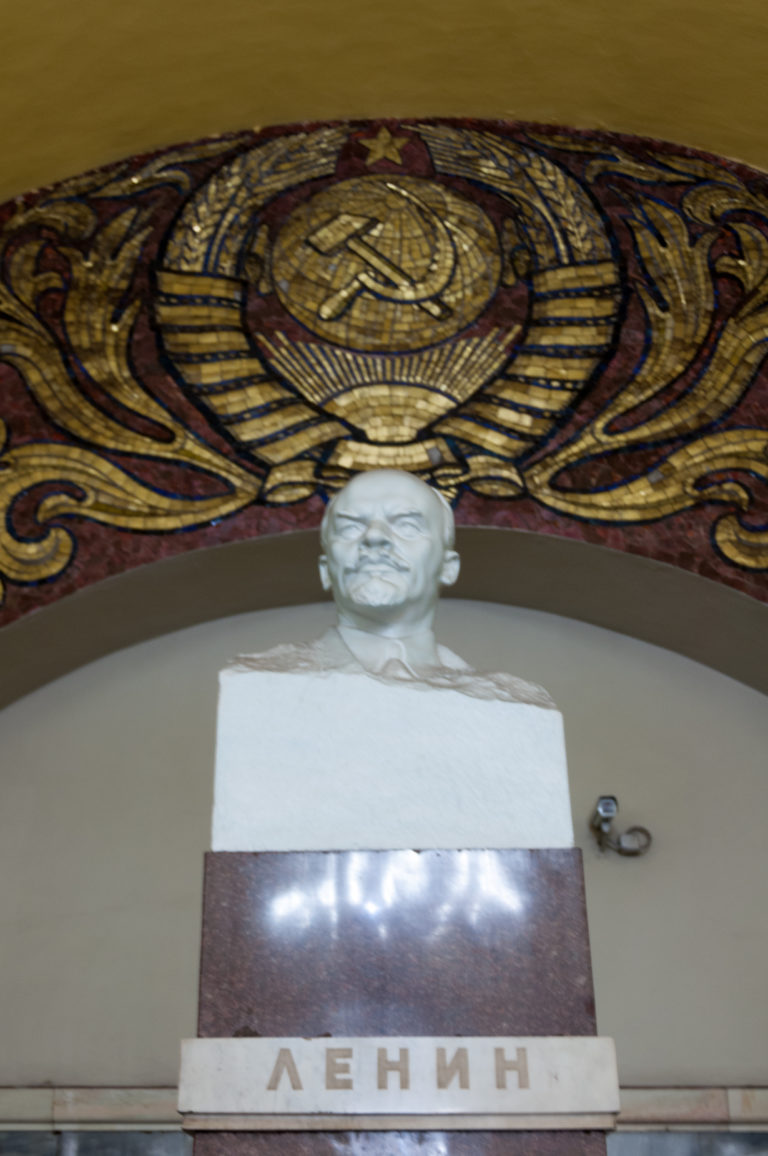 White bust of Vladimir Lenin on a pedestal labeled ЛЕНИН beneath a large hammer-and-sickle mosaic emblem.