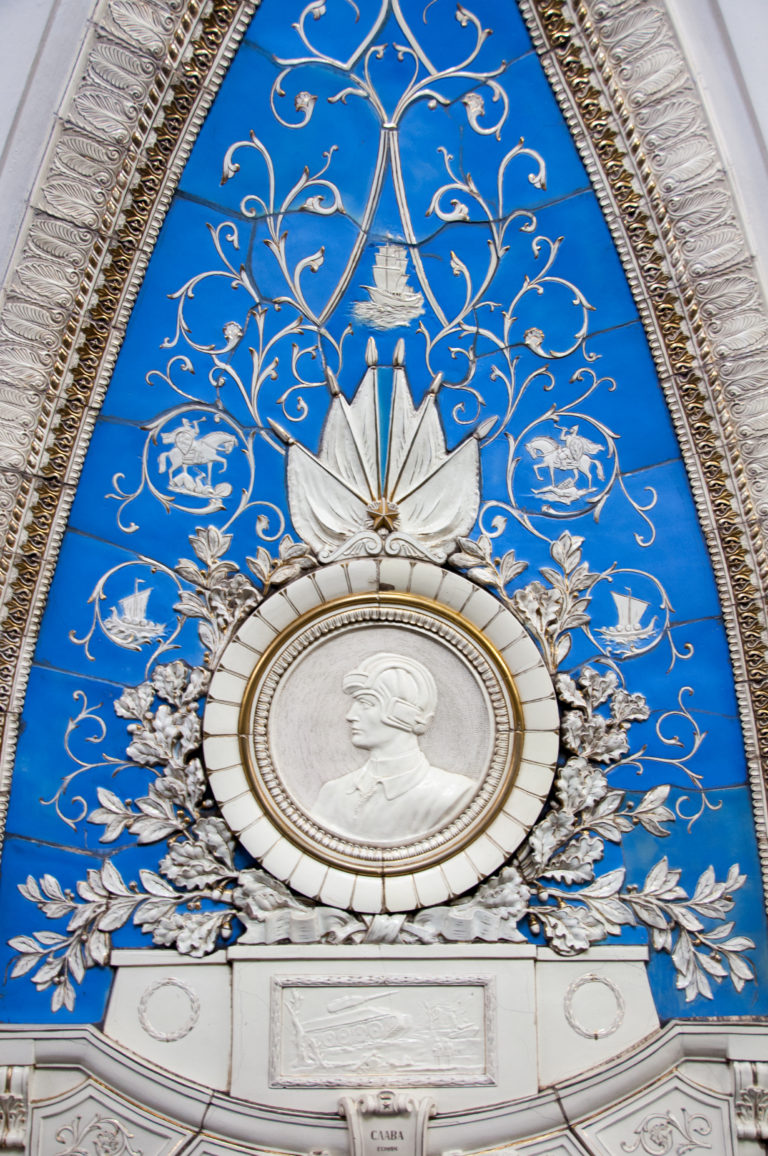 White profile portrait medallion centered on a bright blue panel with pale floral scrolls, ribbons, and relief ornament.