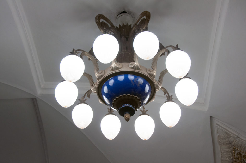 Large chandelier with ring of white globes, dark metal arms, and a deep blue central bowl against a pale ceiling.