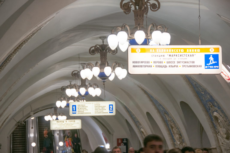 Passengers beneath chandeliers and Cyrillic directional signs in a white-vaulted Moscow Metro passage with decorative side arches.