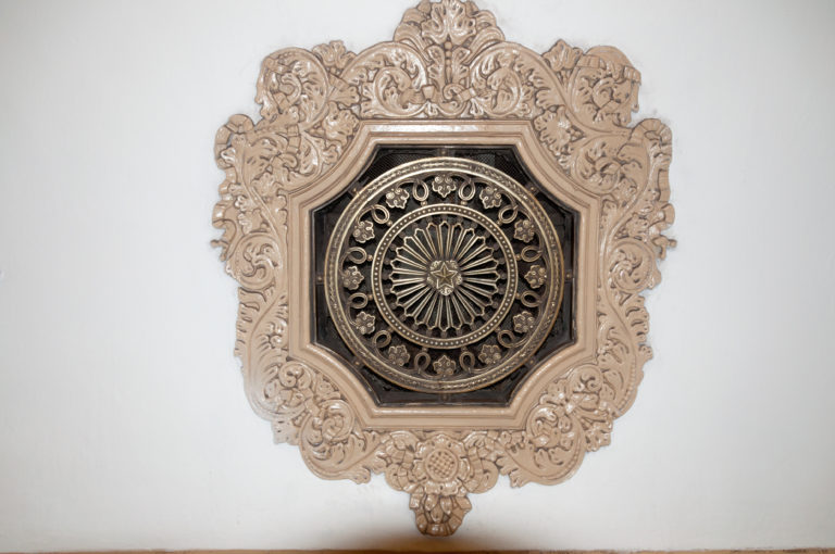 Octagonal metal ventilation grille with radial pattern inside an elaborate cream plaster frame of scrolls and floral ornament.