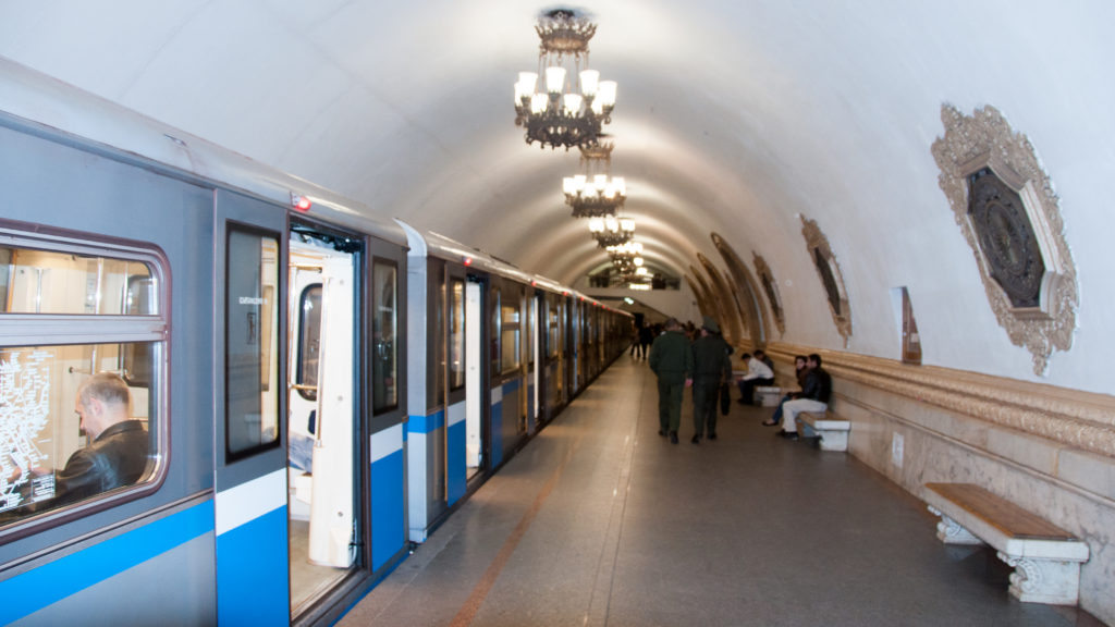 Blue-and-silver metro train beside a chandelier-lit vaulted platform with passengers, benches, and ornate wall decoration.