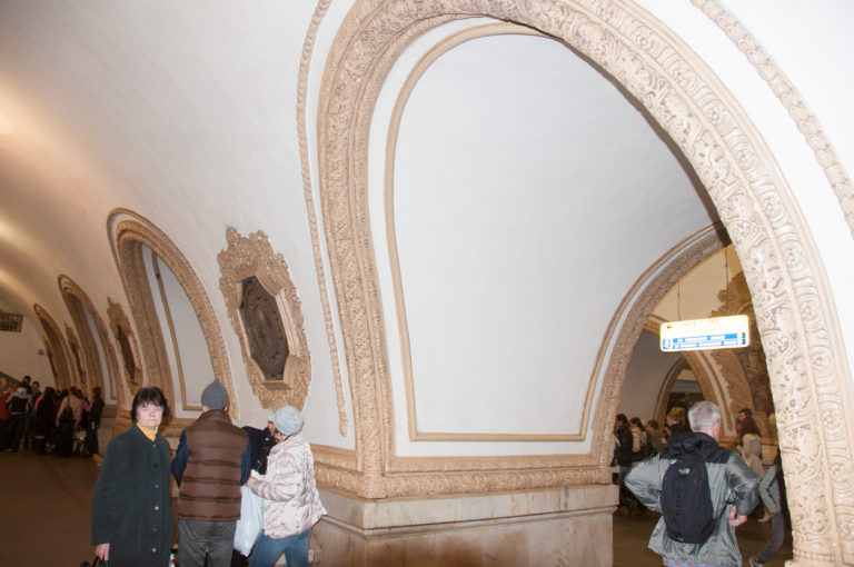 Large white metro arch with layered carved border and stone base, framing passengers and an adjoining decorated hall.