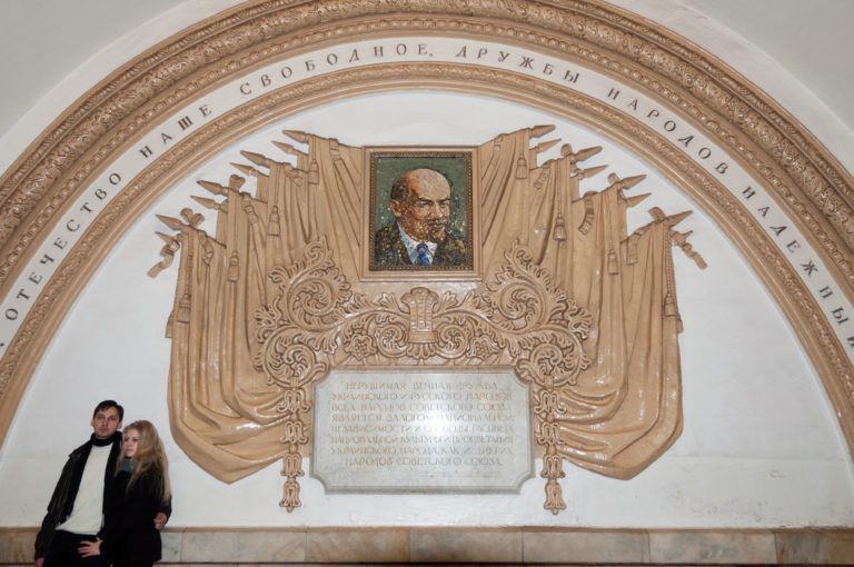 Portrait of Vladimir Lenin above a large Cyrillic inscription, banners, and ornate Soviet wall decoration; two passengers sit below.