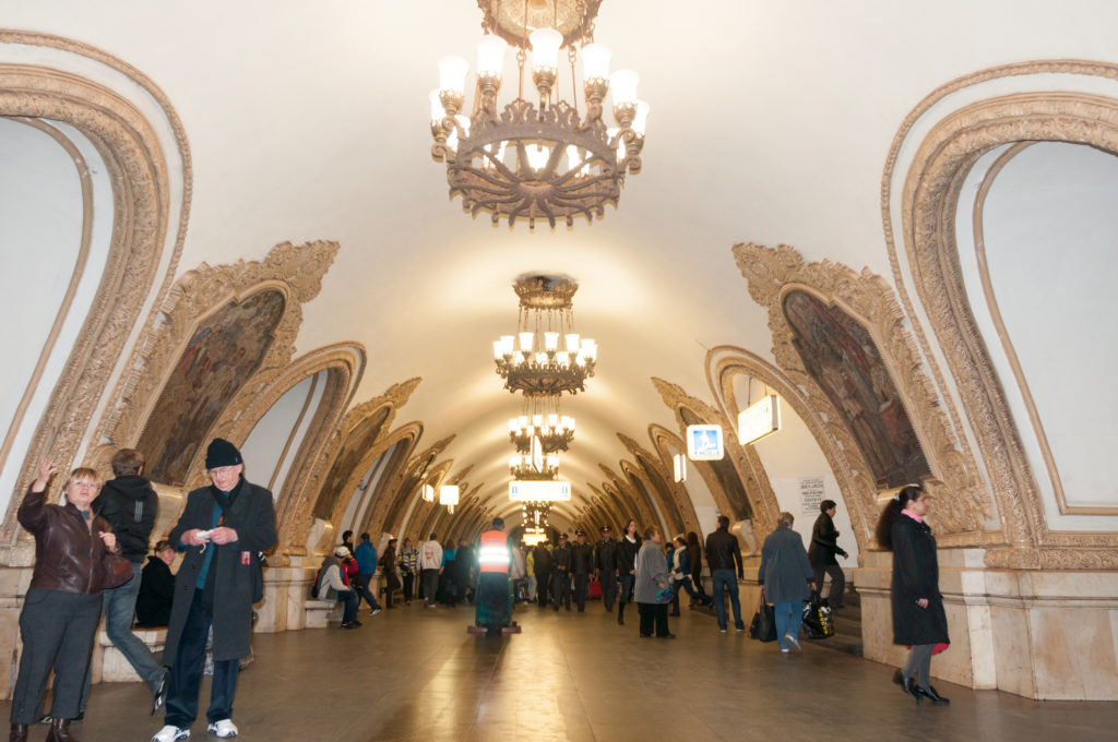 Long symmetrical metro hall with chandeliers, ornate framed wall art, and passengers spread across the central floor.