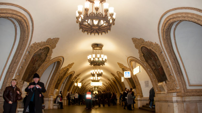 Passengers beneath large chandeliers in a vaulted metro hall lined with ornate arched mosaic frames.