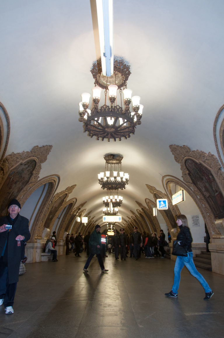 Wide vaulted Moscow Metro hall with ornate chandeliers, framed wall mosaics, and passengers crossing the floor.