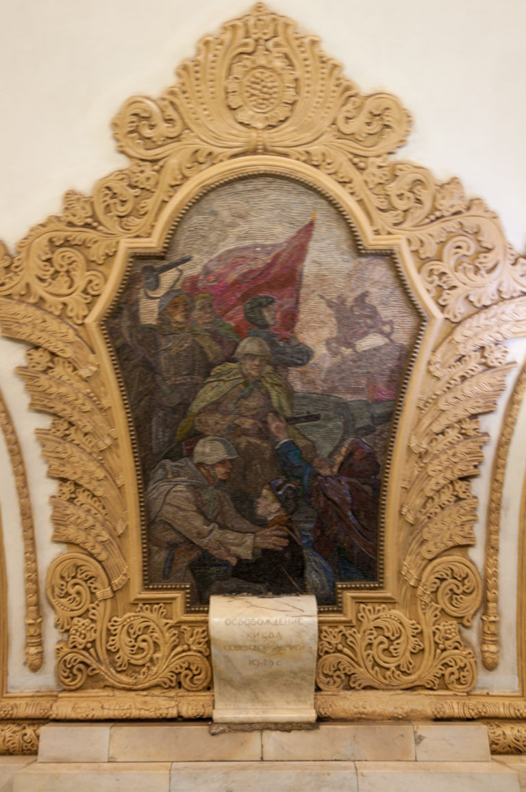 Mosaic of soldiers beneath a red flag set inside an ornate carved frame on a Moscow Metro wall.