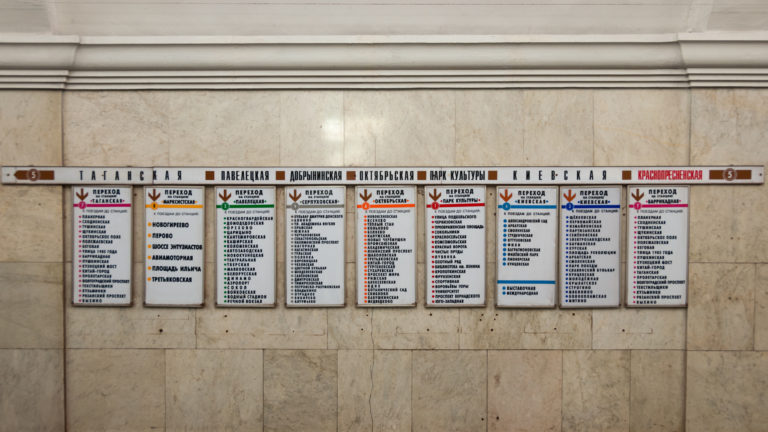 Row of Cyrillic Moscow Metro direction boards with colored arrows mounted across a pale marble wall.
