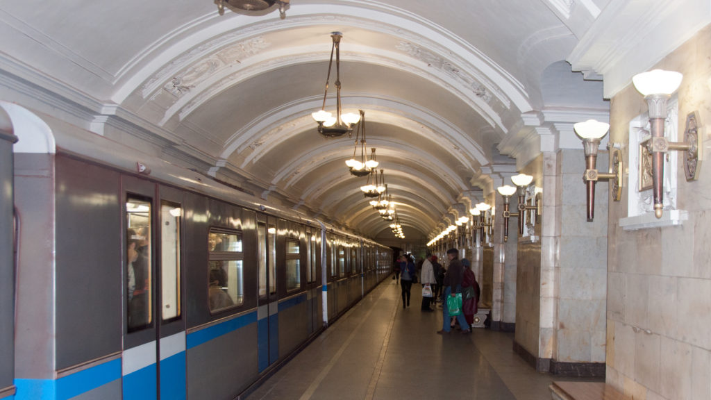 Silver metro train with blue stripe standing beside a long vaulted platform with passengers walking in the distance.