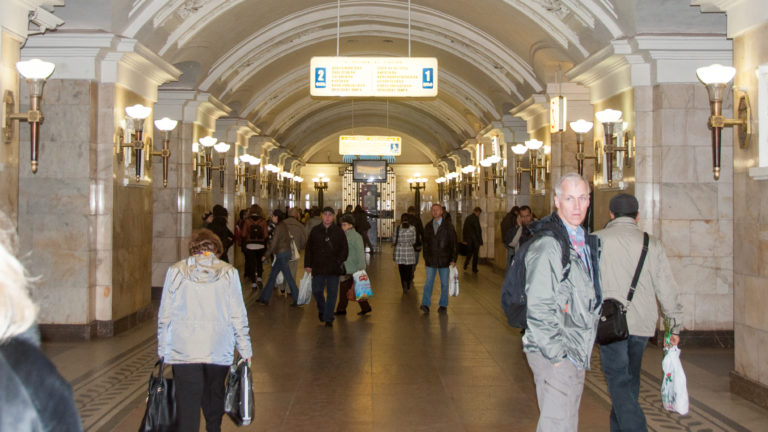 Crowded marble-lined Moscow Metro passage with repeated arches, wall lamps, and overhead directional signs.