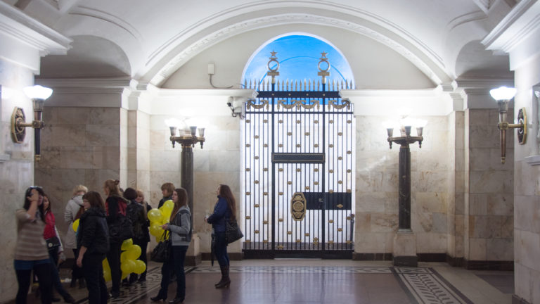 Ornamental metal gate under a blue-lit arch, flanked by wall lamps, with passengers and yellow balloons at left.
