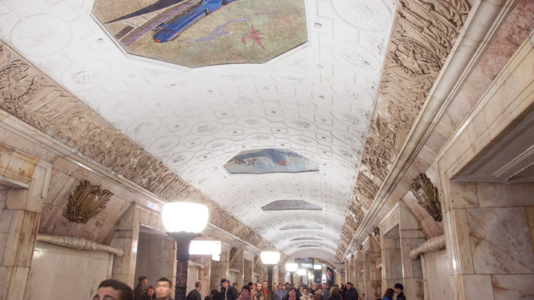 Upward view along a white coffered metro vault with colored mosaics and sculpted relief bands above passengers.