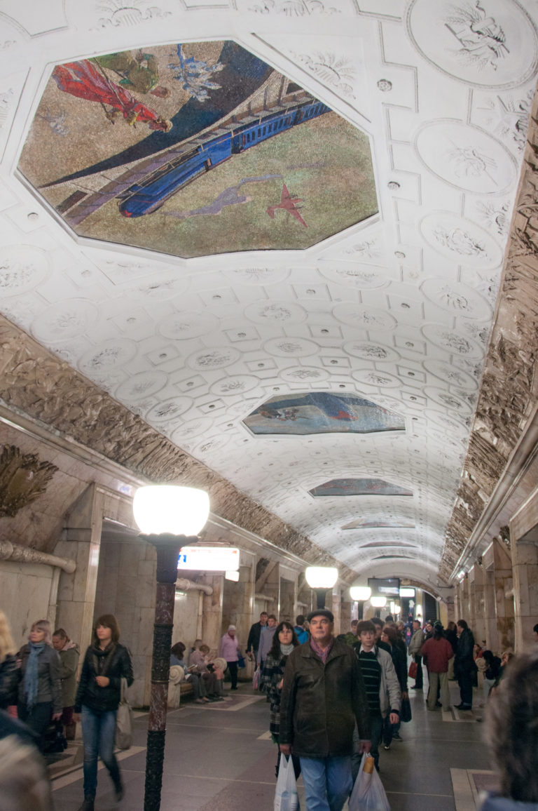 Passengers walking through a metro hall beneath ceiling mosaics, white coffers, relief friezes, and dark lamp standards.