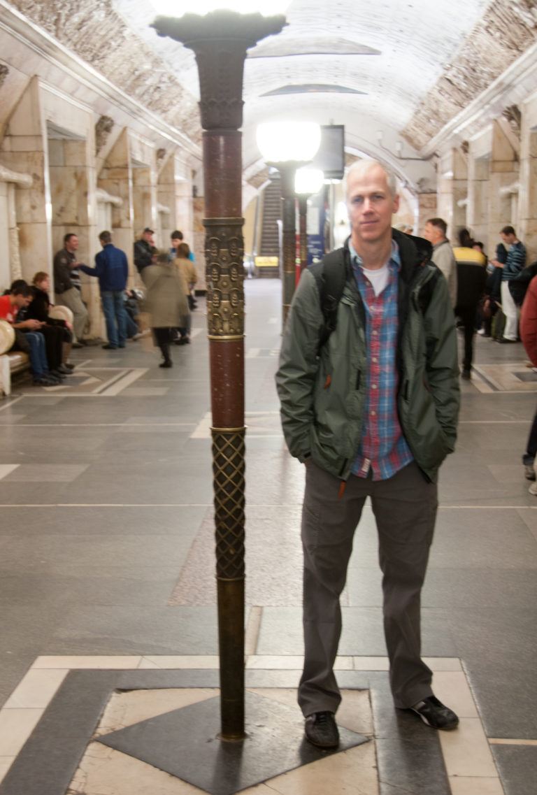 Man standing beside a dark lamp standard in a decorated Moscow Metro hall with seated passengers behind.