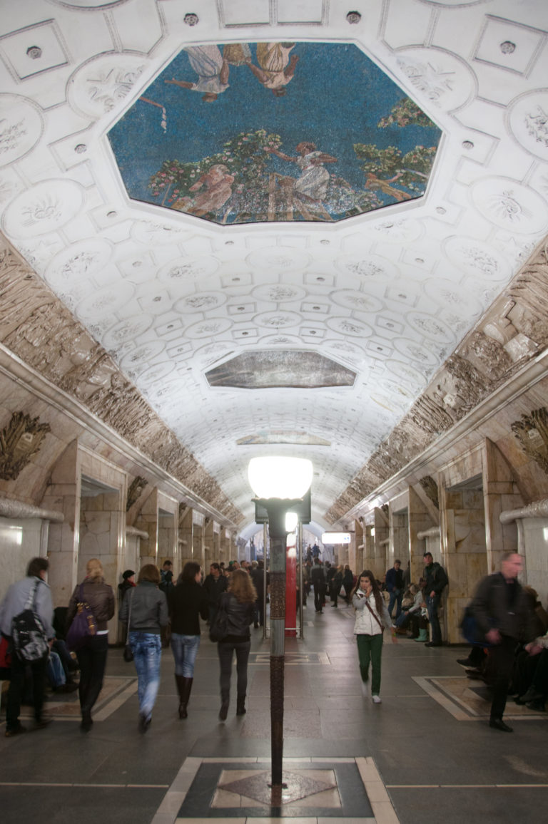 Long metro hall with white coffered ceiling, colorful mosaic panels, sculptural friezes, lamps, and passengers.
