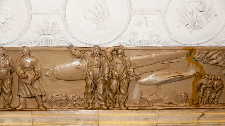 Tan bas-relief showing uniformed figures, civilians, and a large aircraft beneath decorative ceiling medallions.