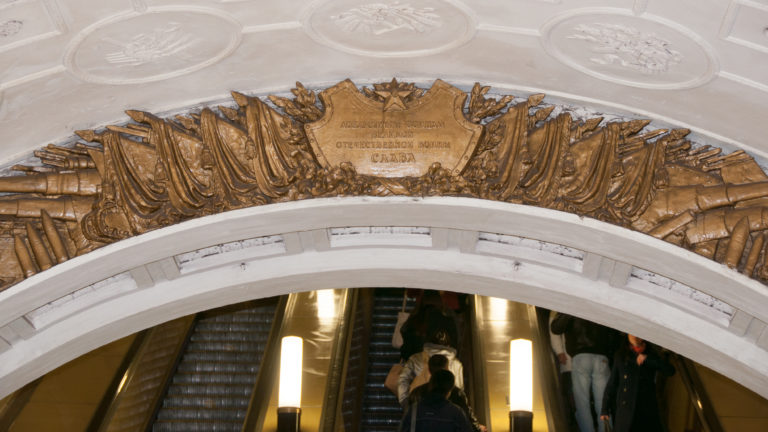 Gilded Soviet-style relief and central inscription above a Moscow Metro escalator opening with passengers below.
