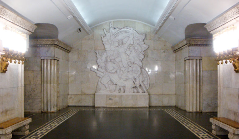 Large pale stone figural relief centered between fluted piers in a marble-clad metro interior.