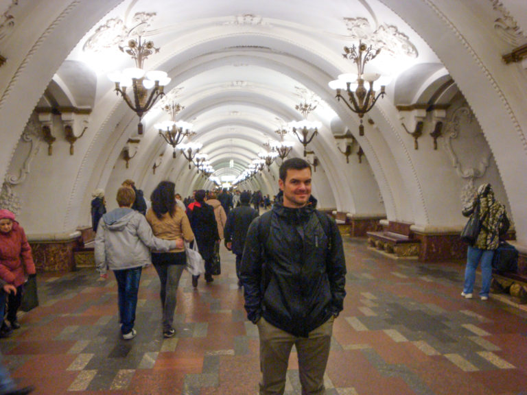 Passengers walk through a long white-vaulted Moscow Metro hall; a man stands at right foreground.