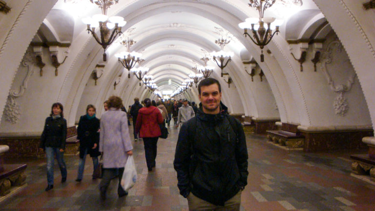 Crowded vaulted Moscow Metro hall with chandeliers, benches, and a man standing in the foreground.