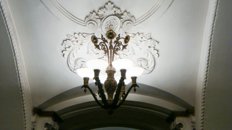 Ornate dark metal chandelier with white glass shades against sculpted white plasterwork above a metro arch.