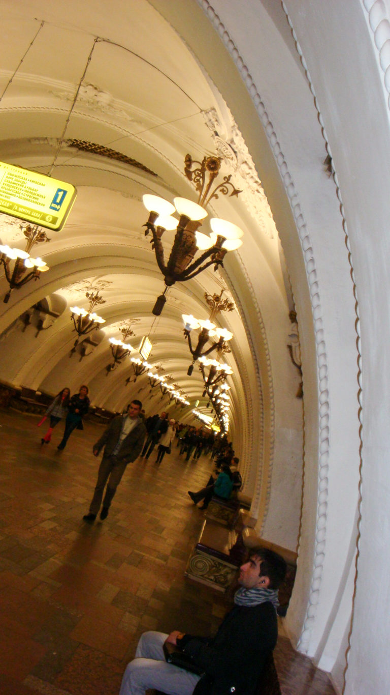 Passengers in a Moscow Metro hall beneath repeating white arches and dark multi-lamp chandeliers.