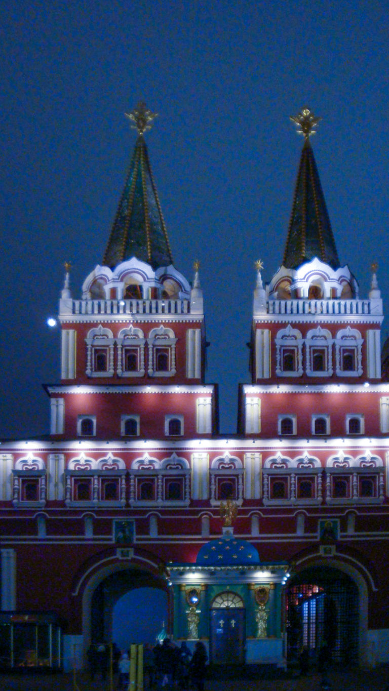 Illuminated twin red-and-white towers of the Resurrection Gate with steep green roofs and arched passages below.