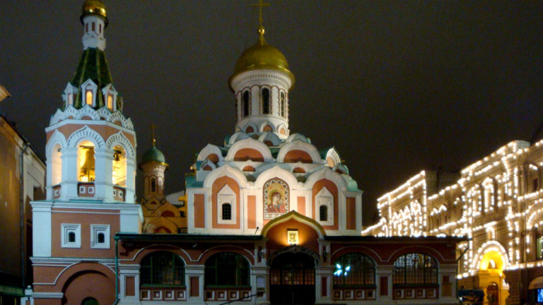 Illuminated red-and-white Kazan Cathedral with gilded dome and bell tower, with GUM lights extending at right.
