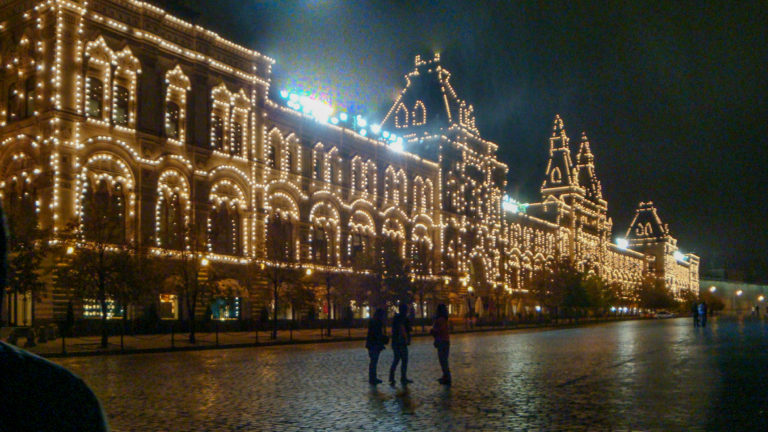 Long illuminated GUM façade beside wet Red Square cobblestones with several pedestrians walking at night.