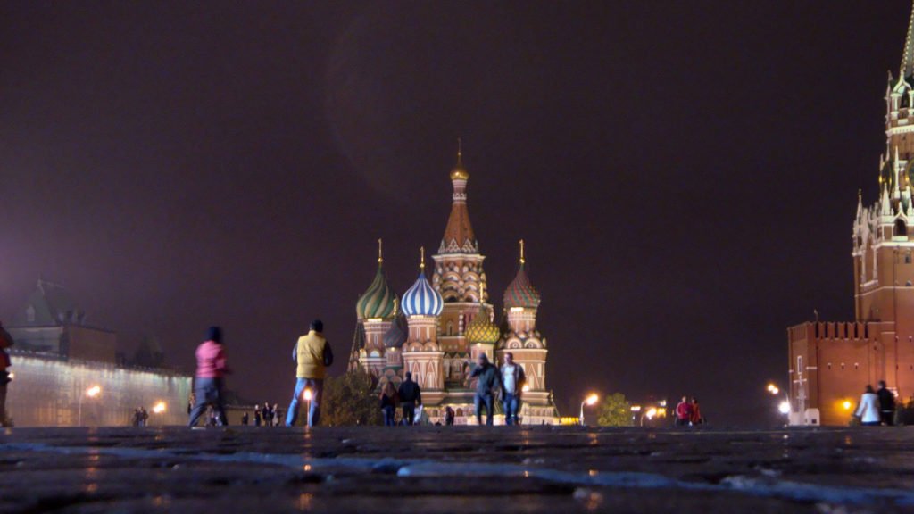 Pedestrians cross wet Red Square cobblestones toward Saint Basil’s Cathedral at night, with the Kremlin at right.