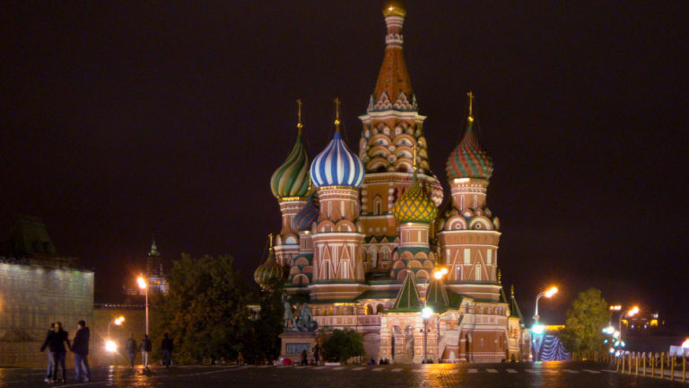 Frontal nighttime view of illuminated Saint Basil’s Cathedral with colorful domes, streetlamps, and a few pedestrians below.