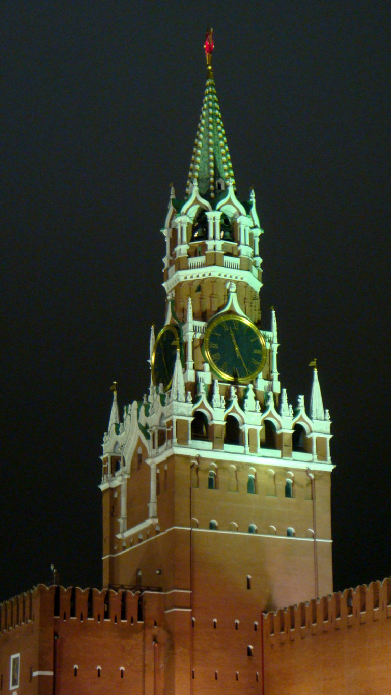 Illuminated Spasskaya Tower with glowing clock, green spire, red star, and crenellated Kremlin wall at night.