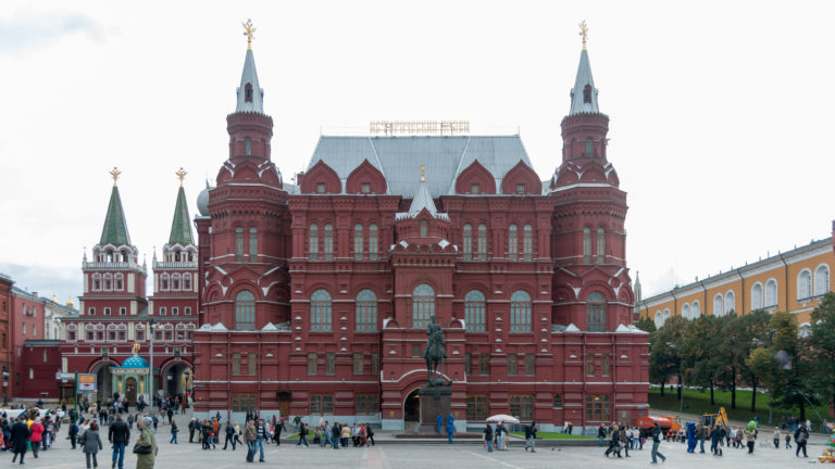 Frontal view of the red-brick State Historical Museum with towers and spires, pedestrians spread across Red Square in front.