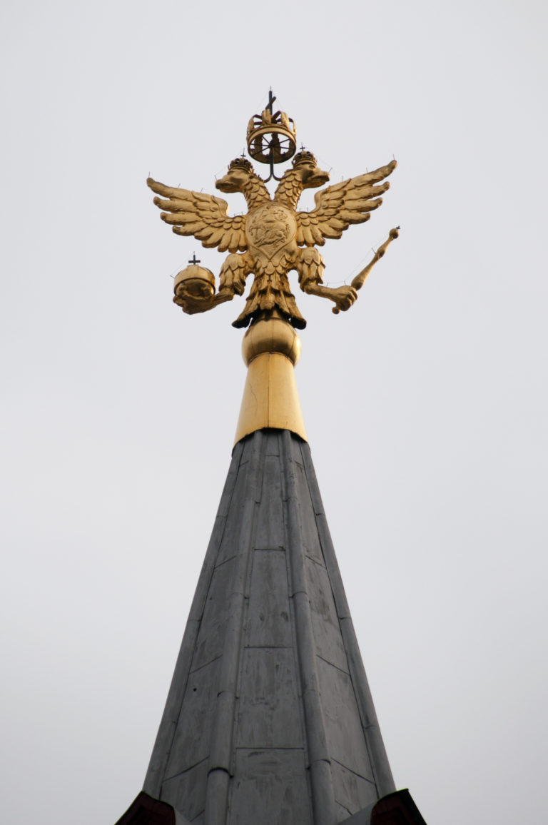 Gilded Russian double-headed eagle with spread wings and crowns mounted at the peak of a gray museum roof.