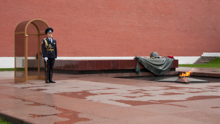 Uniformed honor guard standing beside the Eternal Flame and Tomb of the Unknown Soldier memorial against the Kremlin wall.
