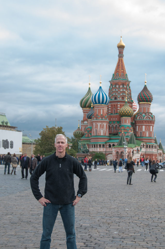 Full-length man standing hands on hips on Red Square, with Saint Basil’s Cathedral and pedestrians behind him.
