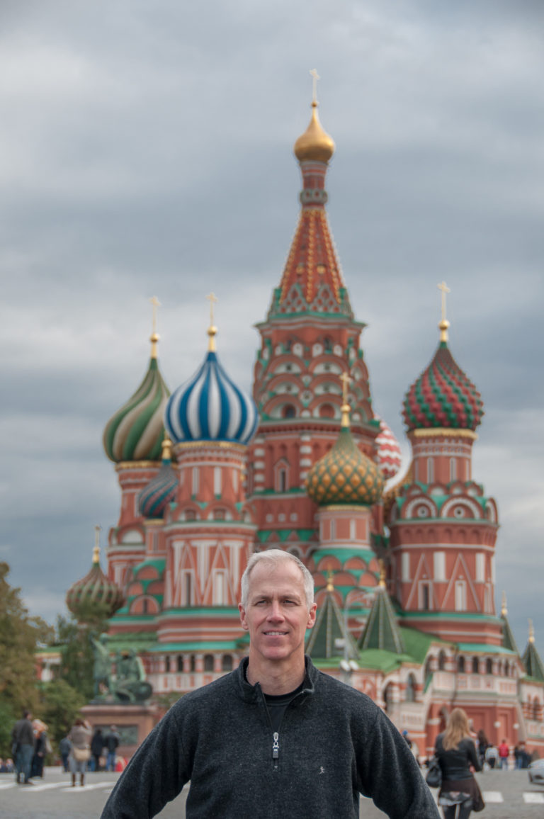 Man standing in front of Saint Basil’s Cathedral, with its colorful domes and central tower softly out of focus.