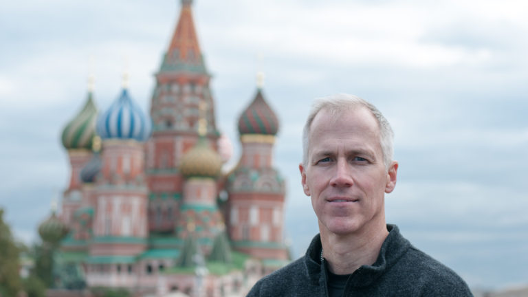 Man facing the camera with the colorful domes and red towers of Saint Basil’s Cathedral blurred behind him.