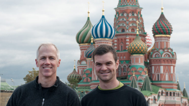 Two men standing side by side with the colorful towers and onion domes of Saint Basil’s Cathedral behind them.
