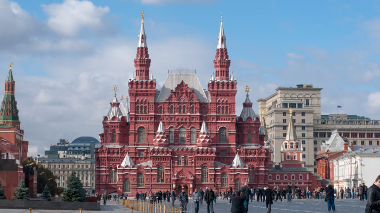 Red-brick State Historical Museum with twin towers at the far end of Red Square, with pedestrians crossing the foreground.