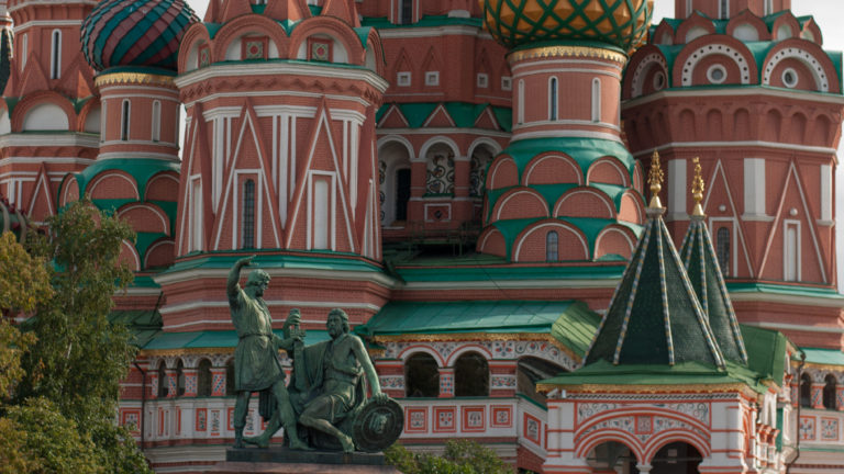 Bronze two-figure monument in front of the ornate red, green, and white lower façade of Saint Basil’s Cathedral.