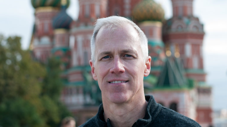 Close portrait of a man in a dark sweater with Saint Basil’s Cathedral blurred behind him.