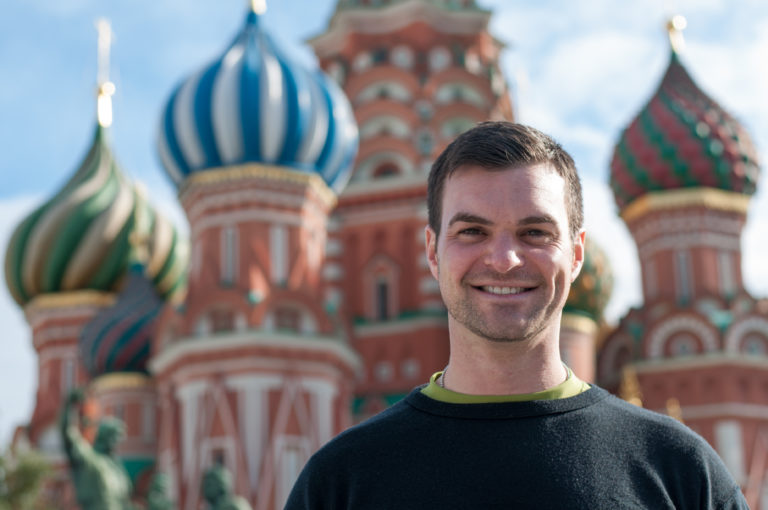 Smiling man in a black sweater centered before the blurred colorful domes and red towers of Saint Basil’s Cathedral.