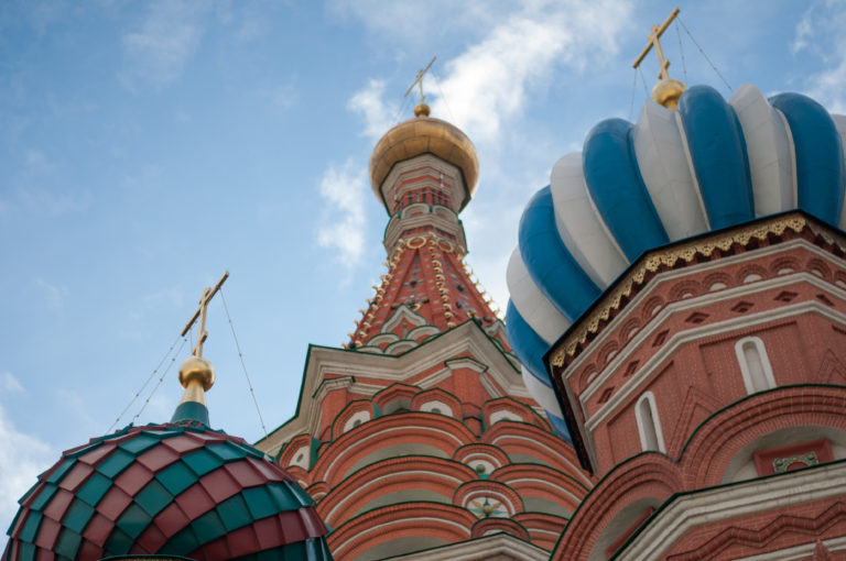 Low-angle close view of Saint Basil’s Cathedral with blue-and-white, green, and gold onion domes above ornate red-brick walls.