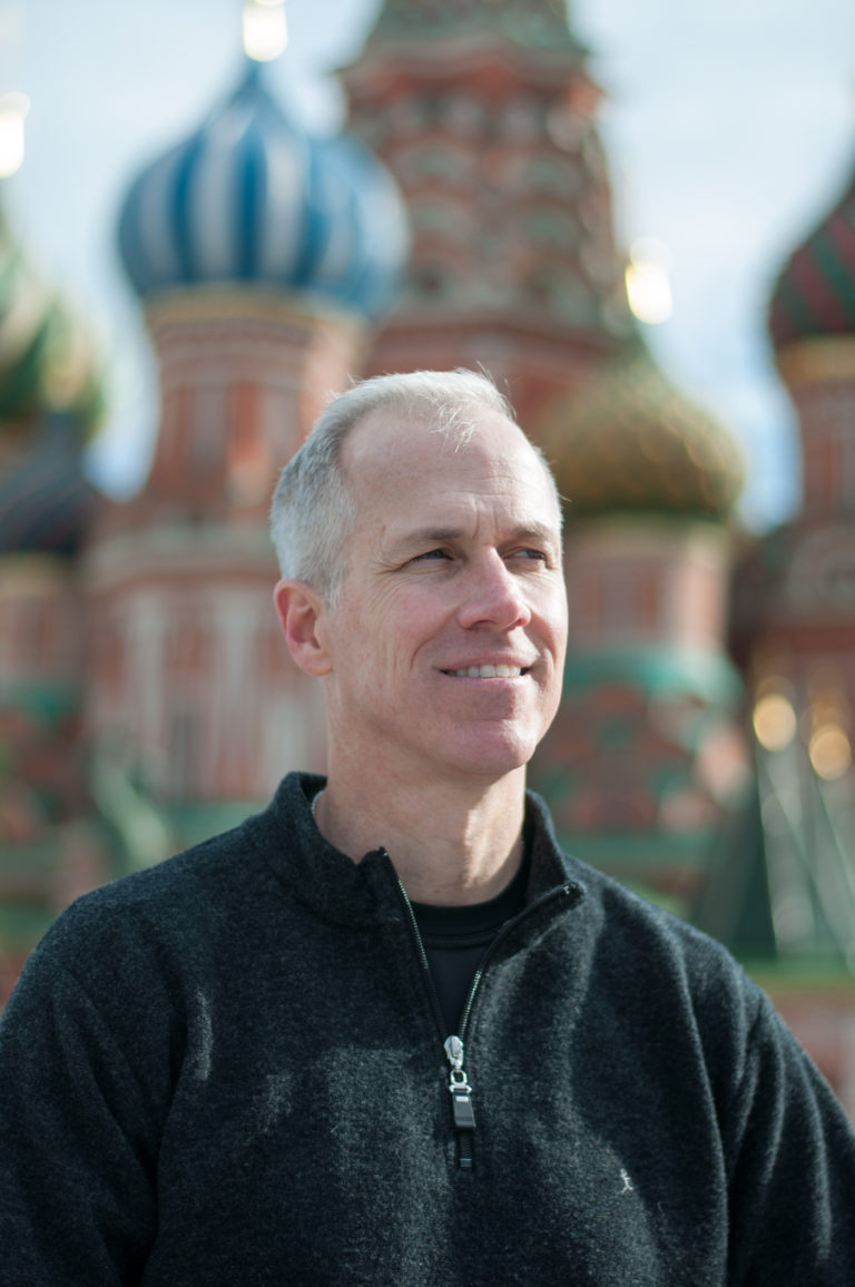 Man in a dark zip-neck sweater looking to the side, with Saint Basil’s Cathedral blurred across the background.
