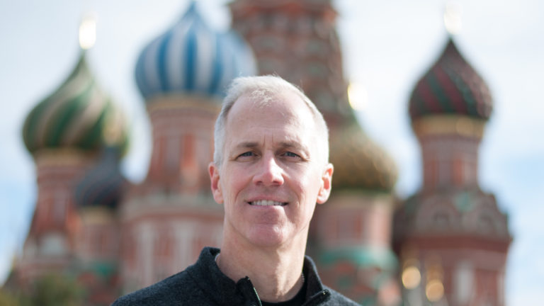 Man in dark sweater facing the camera with the colorful onion domes of Saint Basil’s Cathedral blurred behind him.